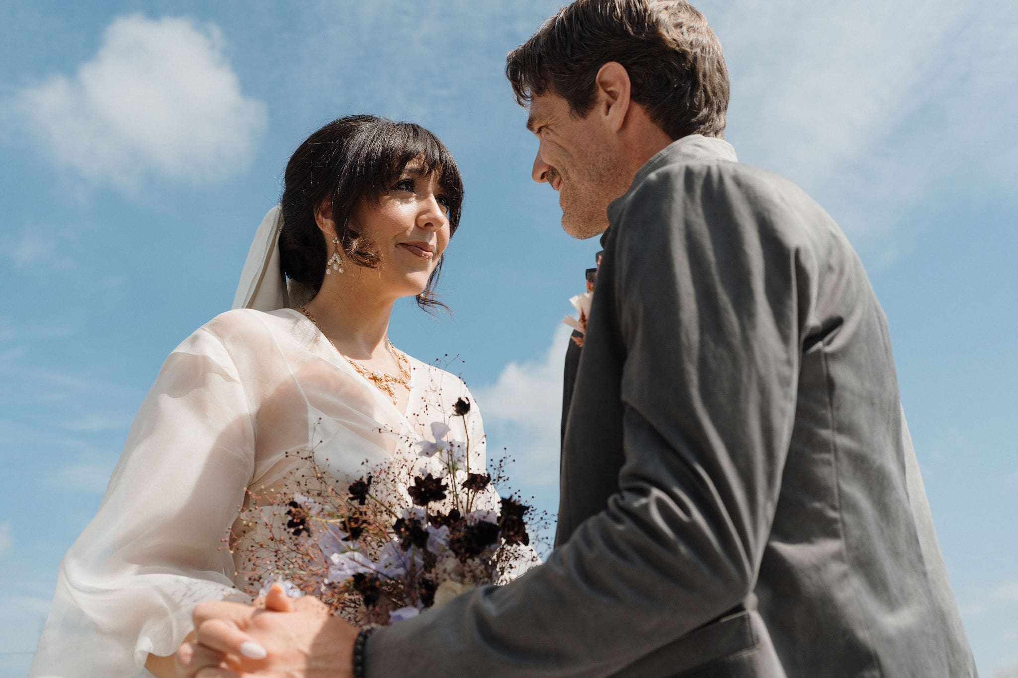 Bride and groom sharing a tender moment against a blue sky, bride holding dark floral bouquet