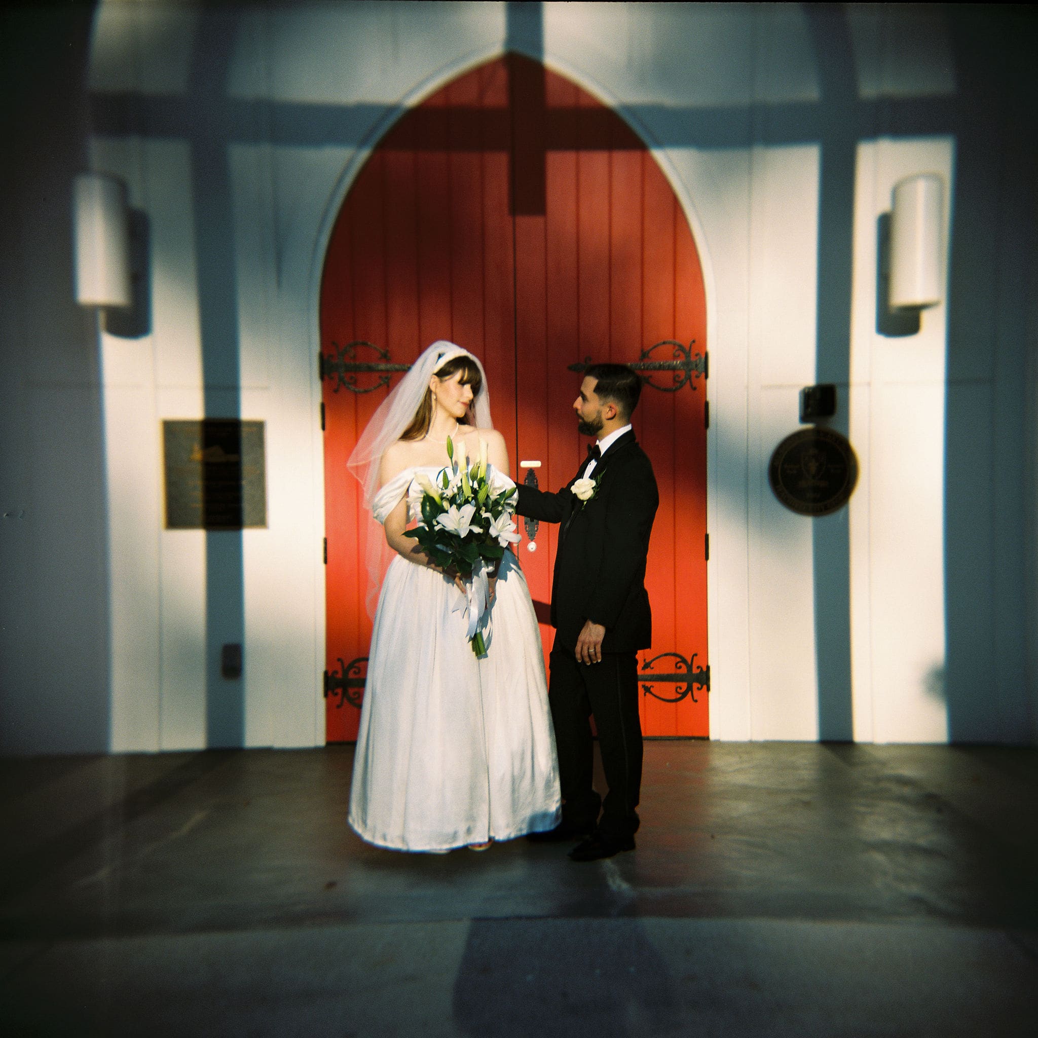 Bride and groom standing before a red arched church door at night, film photography