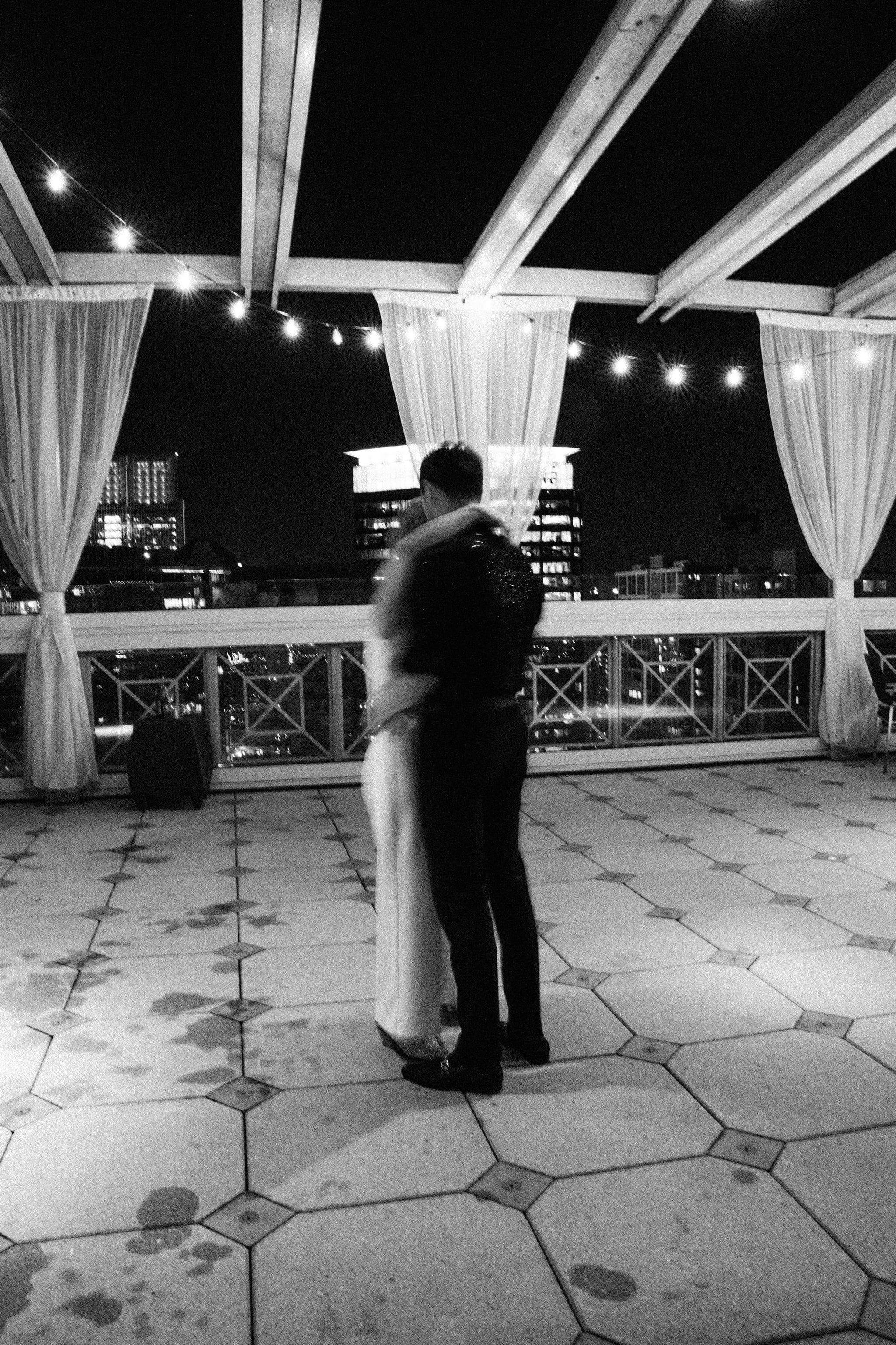 Couple sharing a slow dance on a rooftop terrace with a city skyline at night, black and white