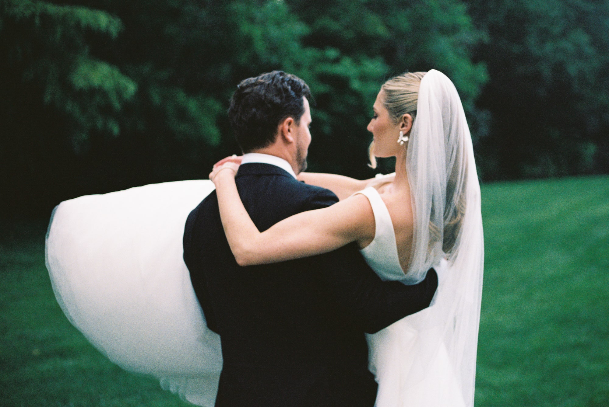 Groom carrying bride in his arms on a lush green lawn, viewed from behind