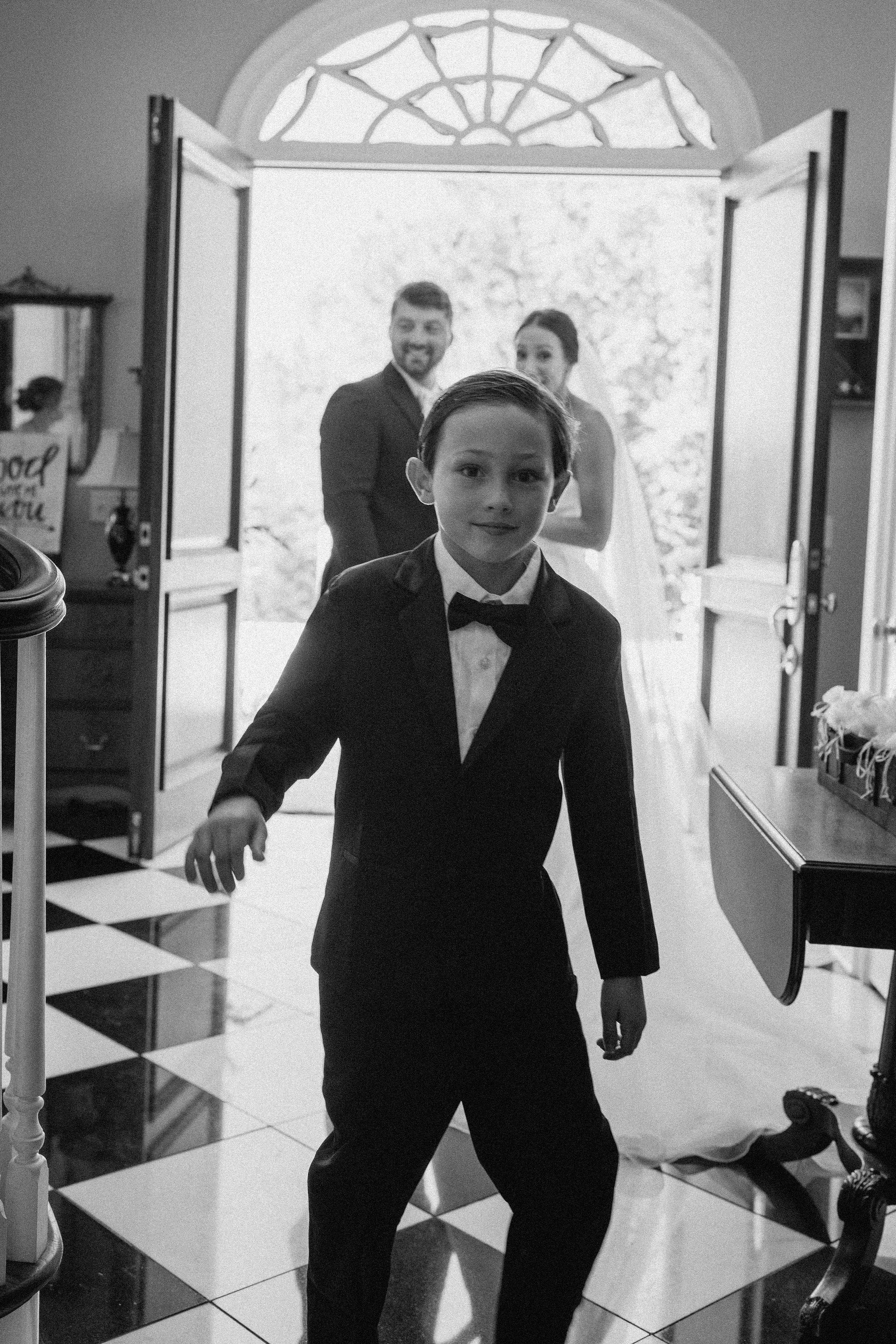 Young ring bearer in tuxedo leading the way as bride and groom arrive, black and white