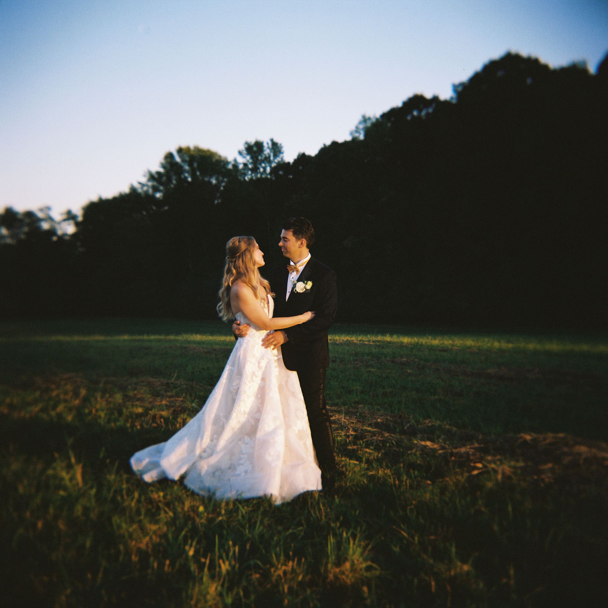 Bride and groom embracing at twilight in an open field with trees silhouetted behind them