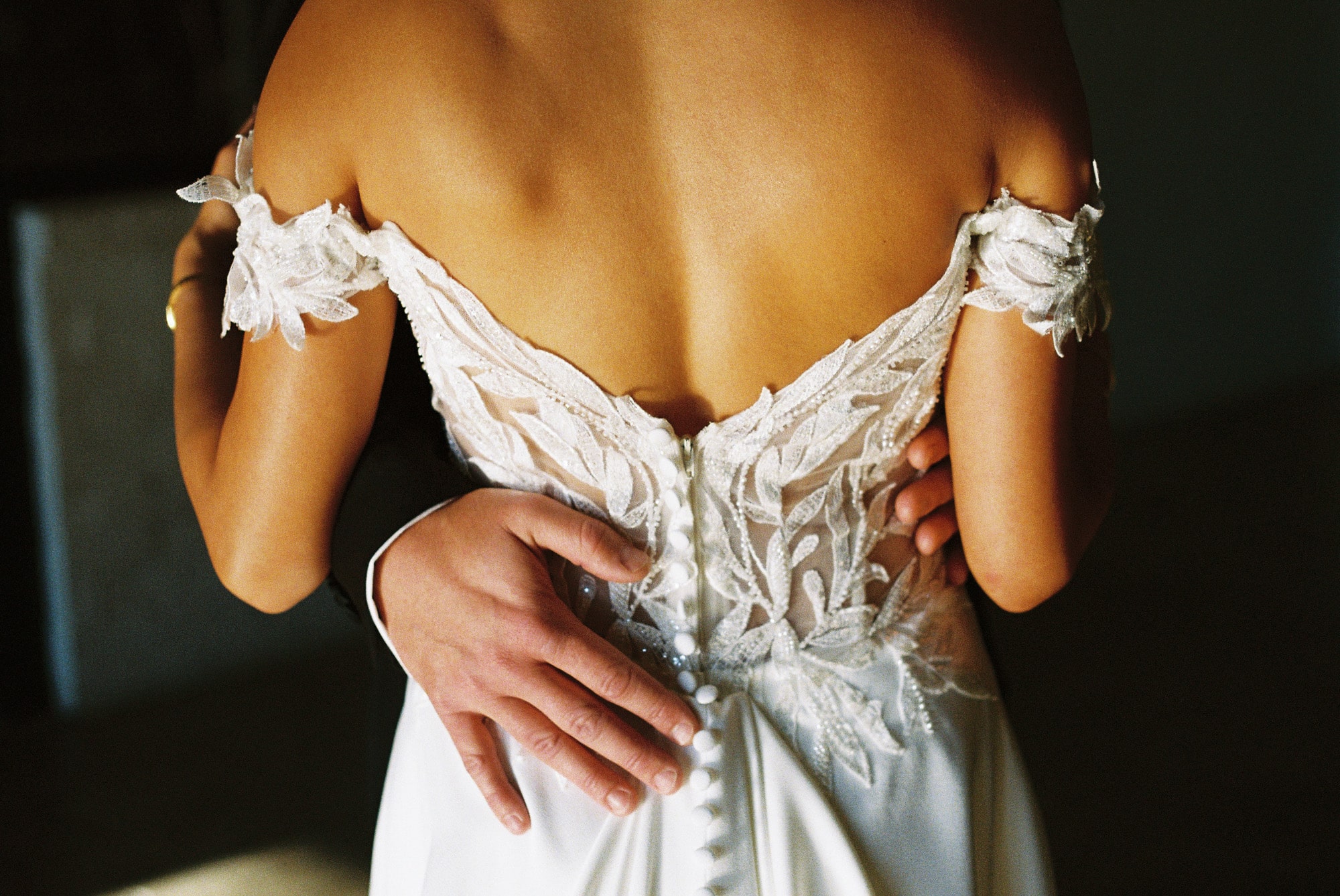 Close-up detail of groom's hands resting on bride's back, highlighting the intricate lace and floral appliqué of her wedding dress