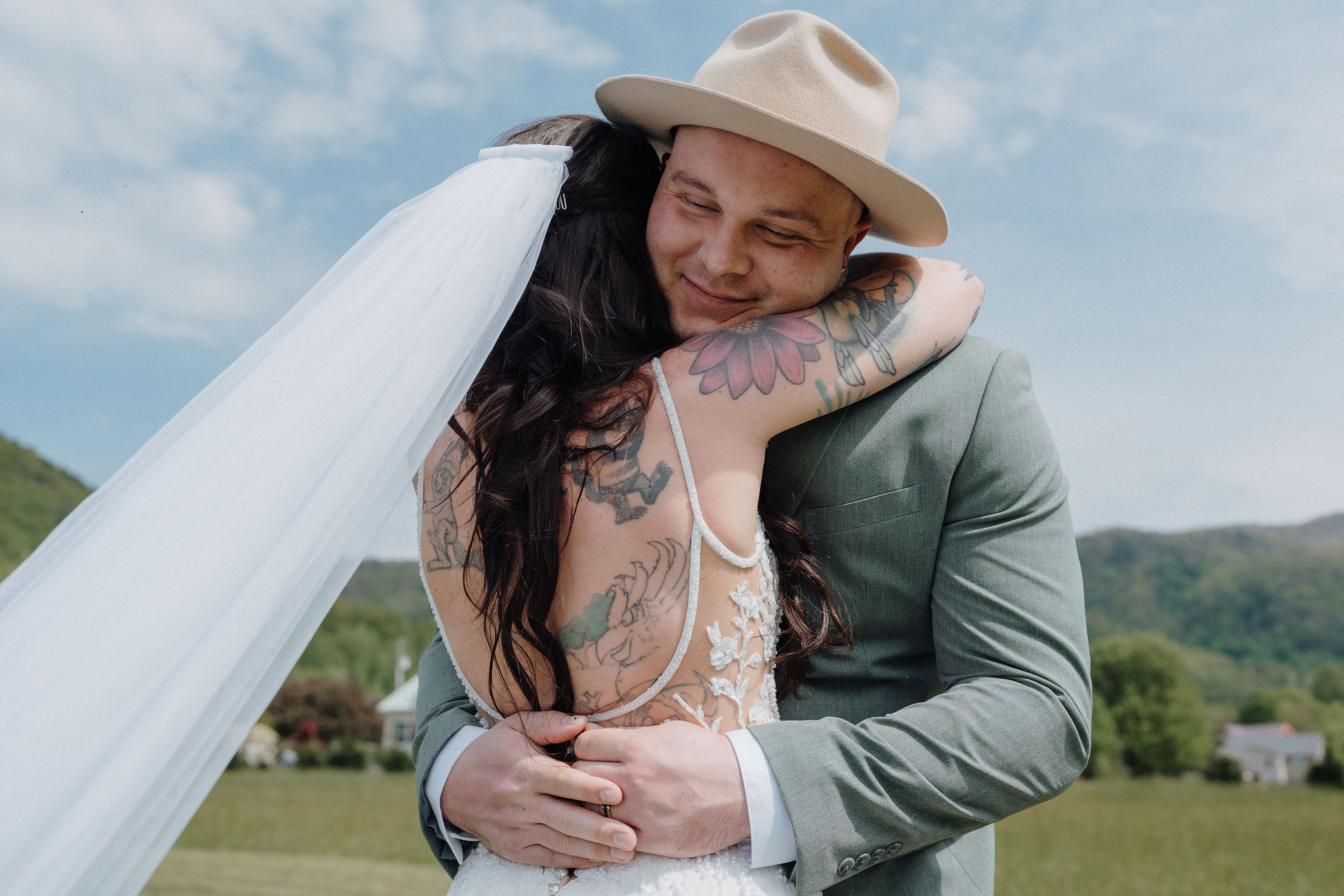 Groom in a tan hat embracing tattooed bride with veil flowing in a mountain meadow